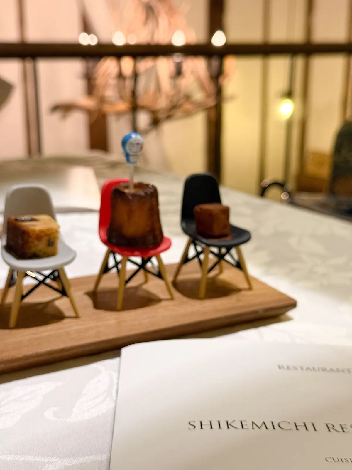 木製のトレーに、3つの小さな椅子にそれぞれ異なる色のケーキが置かれている。背景には、暖色系の照明と木製の柱が見える。下部には「SHIKEMICHI RESTAURANT」と書かれたメニューが置かれている。