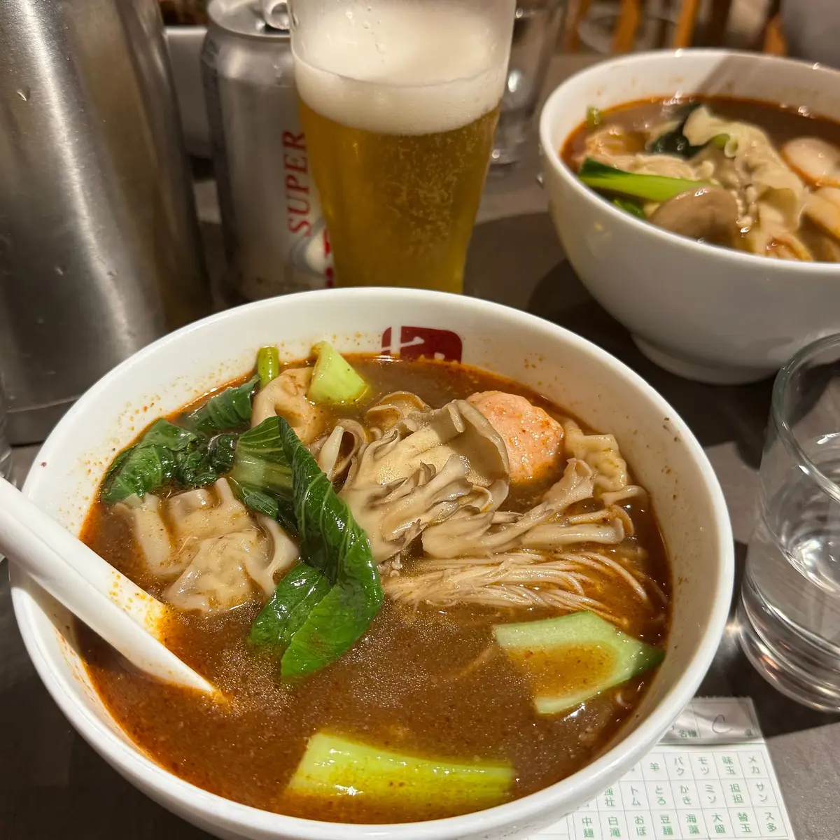 浅めの白い器に入った、赤いスープのラーメン。麺は太めで、具材に豚肉、ネギ、青菜が添えられている。背景にはビールや他の料理が写る。