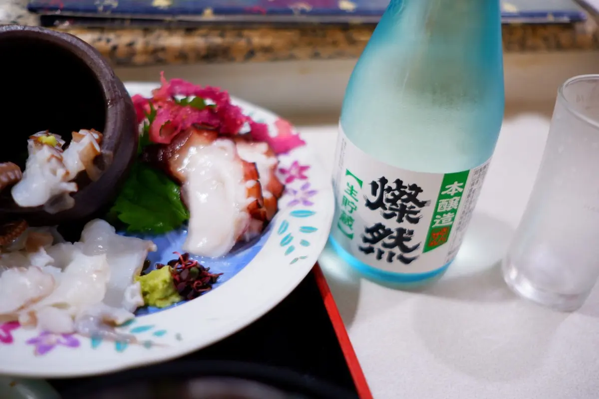 白い皿に盛り付けられたタコ料理と、そばに置かれた「鏡月」と書かれた日本酒のボトル。タコは薄切りでピンク色、皿には薬味とわさびも添えられている。