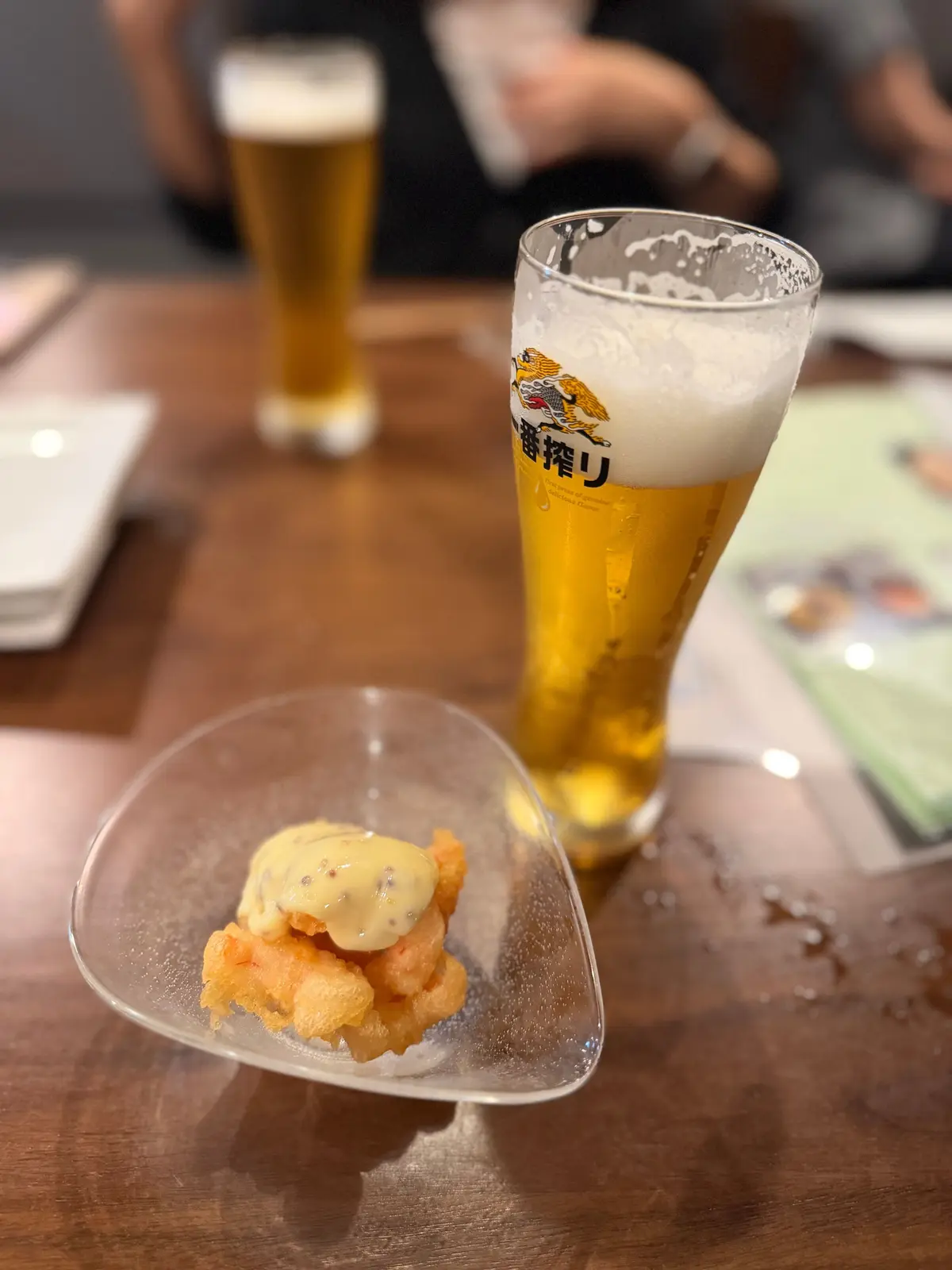 木製のテーブルに置かれたアサヒスーパードライのビールグラスと、ソースをかけた天ぷらを載せた小皿。背景には人物がぼやけて写っている。