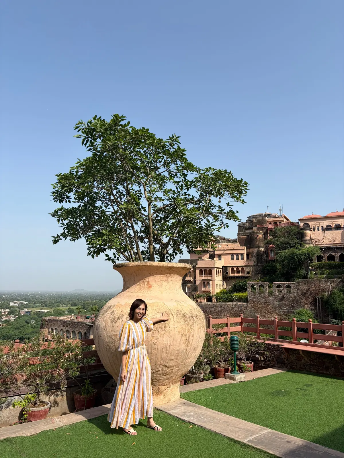 インドの城壁の上の緑の芝生に立つ女性。手前に巨大な壺があり、その中に木が植えられている。背景には、丘の上に建つピンク色の城が見える。