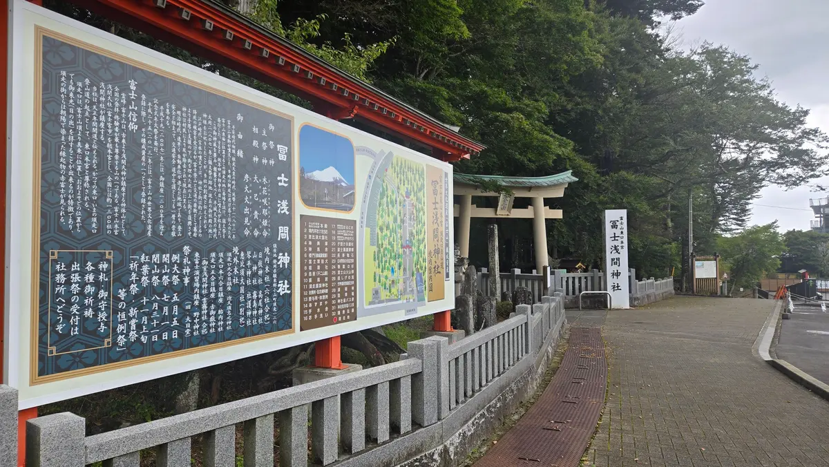 緑豊かな木々に囲まれた鳥居と、その前に設置された緑色の案内板。案内板には漢字で書かれた説明と、山並みのイラストが描かれている。鳥居は朱色で、屋根は緑色。案内板の縁は赤色で装飾されている。