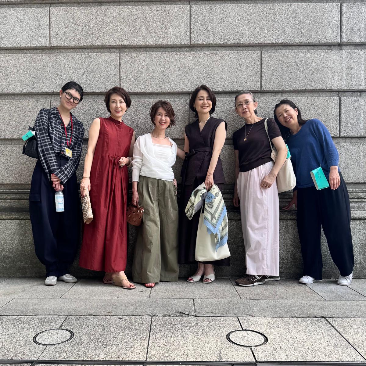 6人の女性が並んで立っている写真。左から、赤いチェックのシャツに黒いパンツ、赤いロングドレス、緑色のパンツに白いトップス、黒いワンピースに水色のスカーフ、黒いカットソーにピンクのパンツ、青いセーターに黒いパンツを着用。背景は灰色の壁。