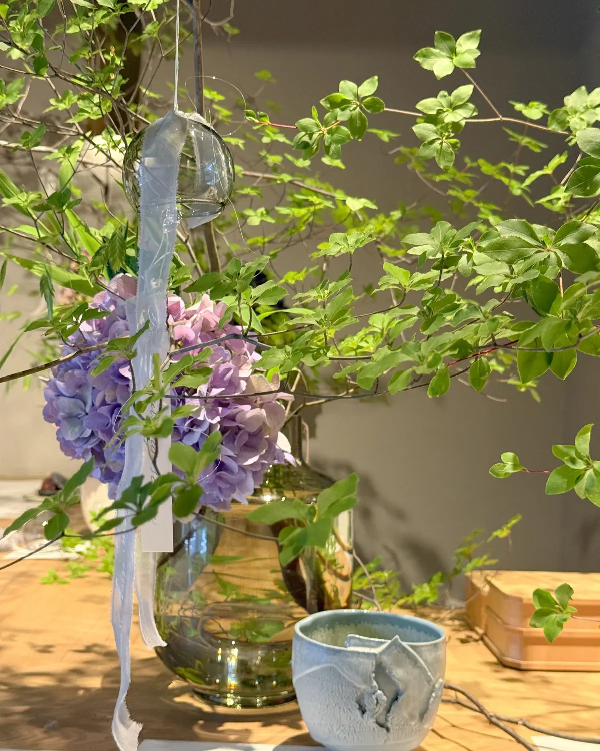 木製テーブルに、緑の葉と紫の花が飾られたアレンジメントと、ガラスの器と白い陶器のコップが置かれています。