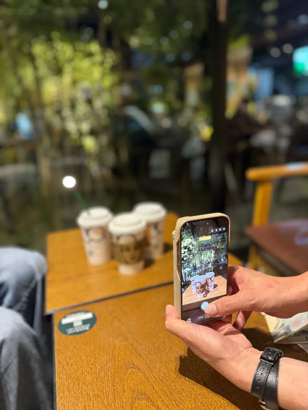 木々に囲まれた屋外のカフェで、スターバックスの紙コップがテーブルに並んでいる。女性がスマートフォンでその様子を撮影している。
