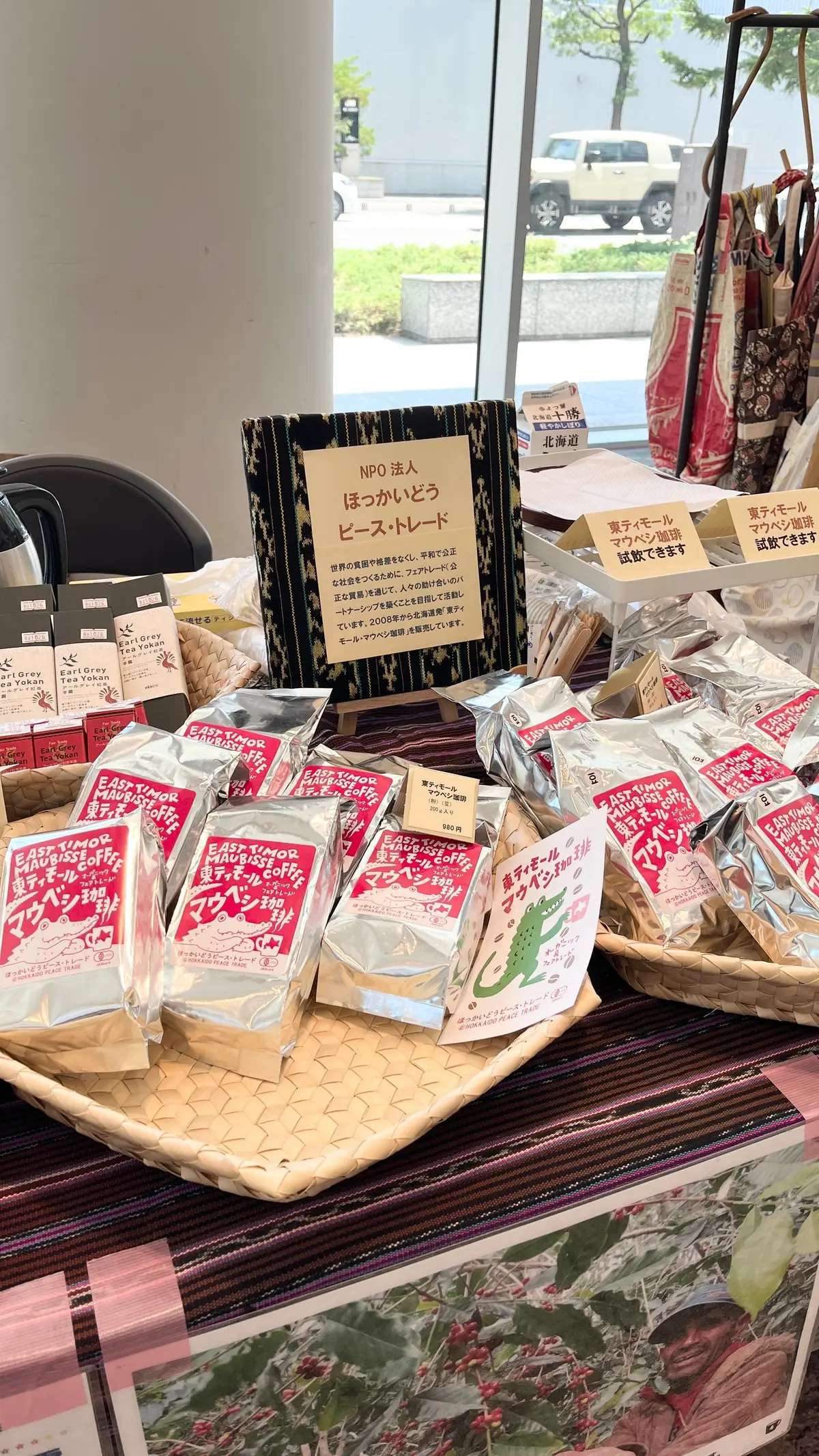 コーヒー豆の小売店内。複数のバスケットに「イーストティモール・モアビッセ・コーヒー」の袋が並び、赤いラベルと緑のワニのデザインが特徴的。店内には窓から外の景色が見え、店員用の椅子やカトラリーも置かれている。
