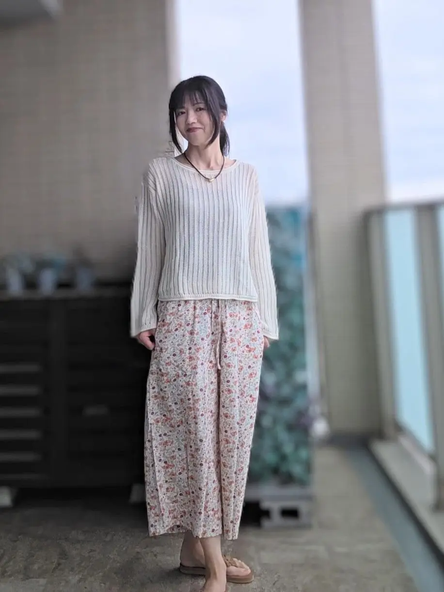 女性がベージュのリブ編みセーターと花柄のロングパンツを着て、屋外のバルコニーに立っています。足元にはサンダルを履いています。背景には白い壁と窓が確認できます。