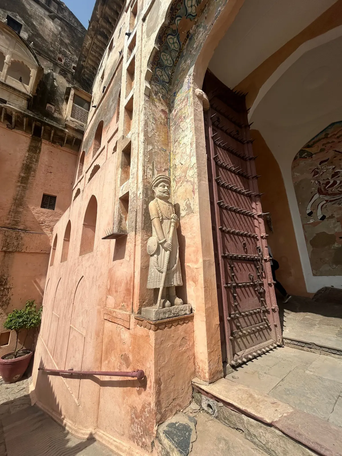 インドの古城の壁面にある彫刻。男性像が杖をつき、壁の角に設置されている。背景にはアーチ型の入り口と、装飾が施された壁が見える。全体的にオレンジ色を基調とした色合い。