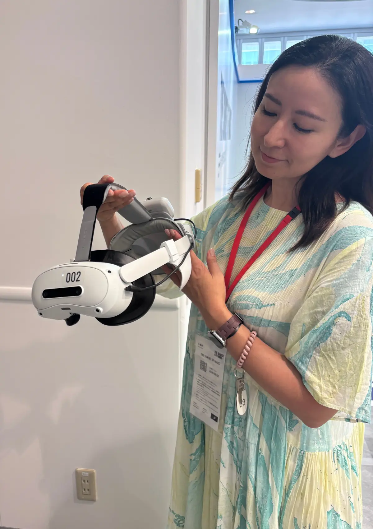 女性が白い360度カメラ型のデバイスを操作している。緑色の葉柄のシャツを着ており、赤いネームタグをつけている。背景には白い壁と窓が見える。