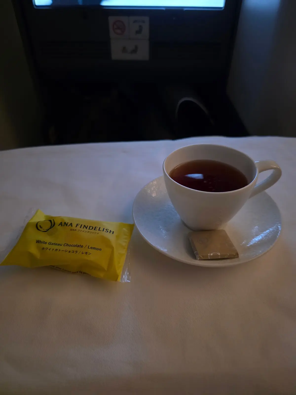 白いテーブルクロスの上に置かれた、ANA Findelishの白い箱とコーヒーカップ。カップには茶色の液体が入っており、ソーサーが添えられている。背景には灰色の座席の背面がある。