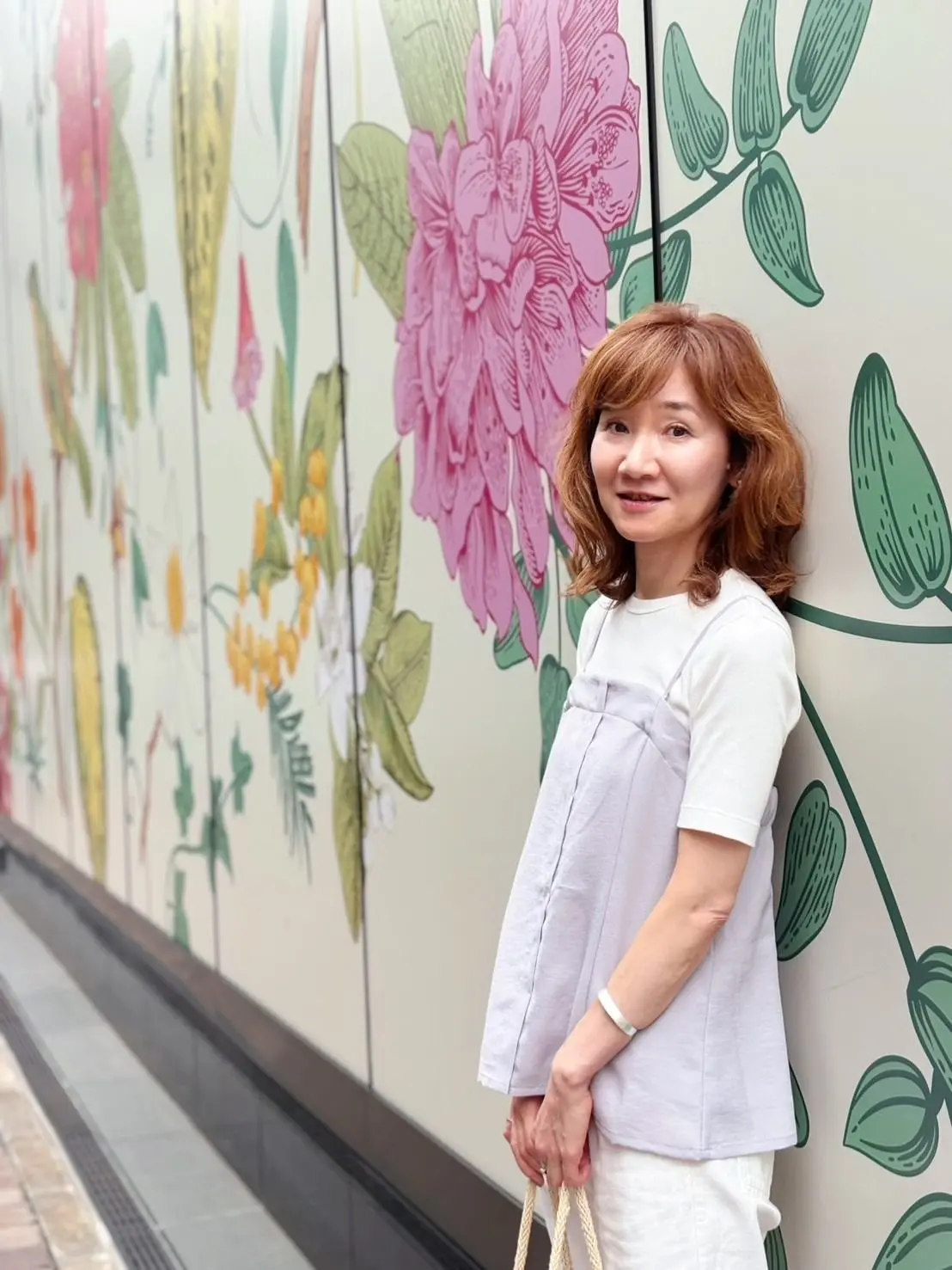 女性が白い壁に寄り添い、花と葉の緑の絵が描かれた背景に立っています。女性は白いトップスとグレーのオーバーオールを着て、笑顔でカメラを見つめています。