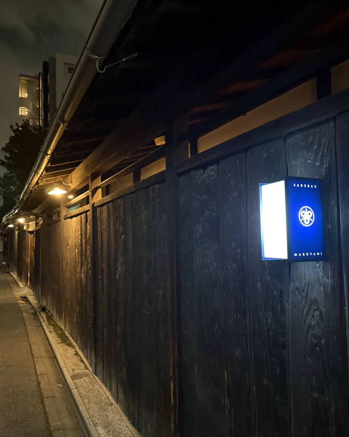 夜の路地裏に沿って続く黒い木製の壁。壁には「SAKEDARU MARUTANI」とある青色の看板が設置されている。壁の照明が灯り、歩道に光が落ちている。