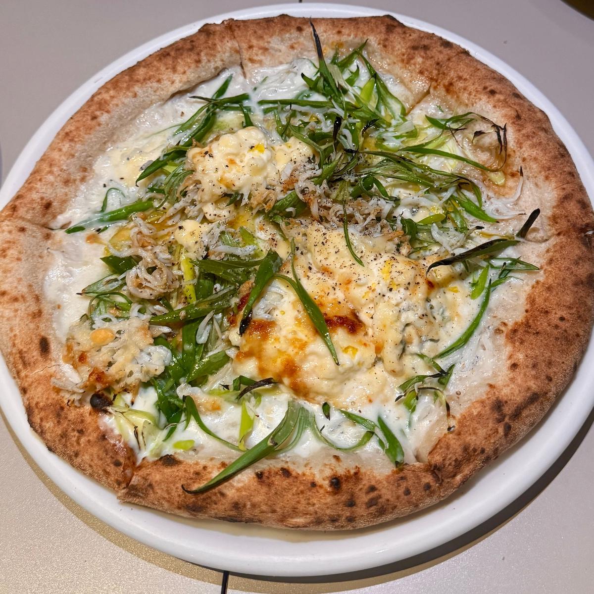 白を基調としたチーズと緑のネギ、リーフレタスが散らされたピザ。縁は焼き色がつき、ふっくらとした生地。白い皿に盛り付けられている。
