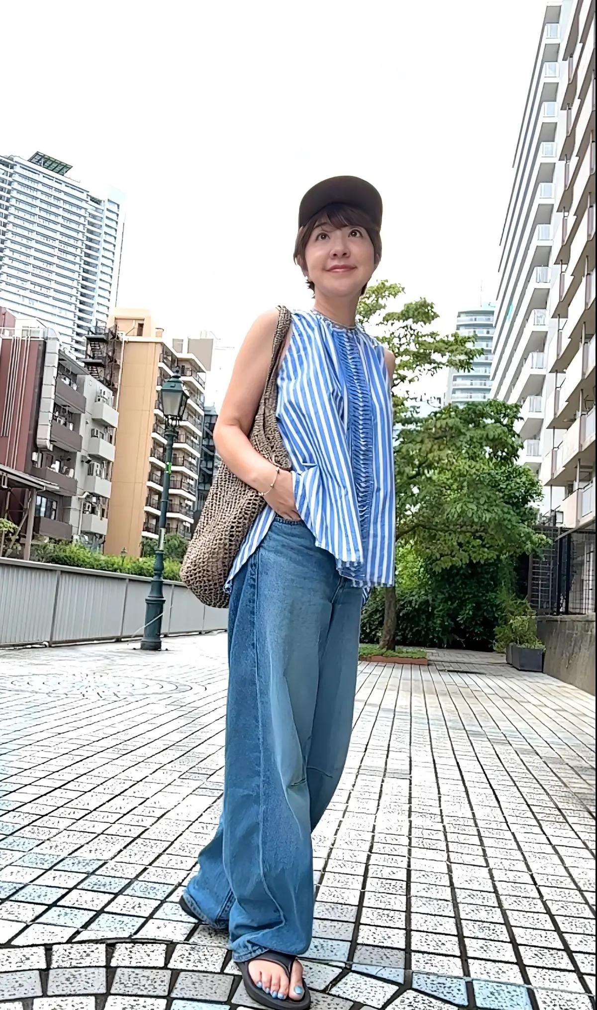 白い帽子を被った女性が、青と白のストライプのトップスとデニムを着用し、笑顔で立っている。背景には高層ビル群と緑の木々が見える。茶色の肩掛けバッグを斜めにかけている。