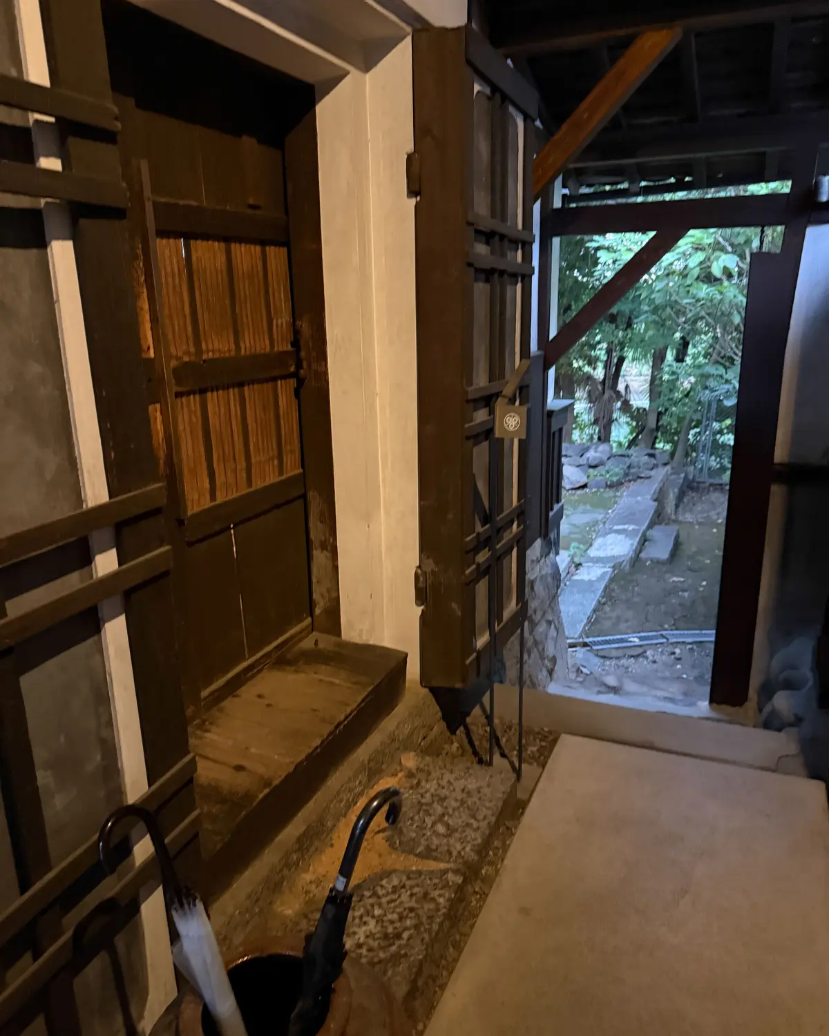 木製の障子戸が二枚開き、緑豊かな庭園が見える。左には洗い場があり、黒い水栓が設置されている。石畳の通路と、木々の緑が目に映る。