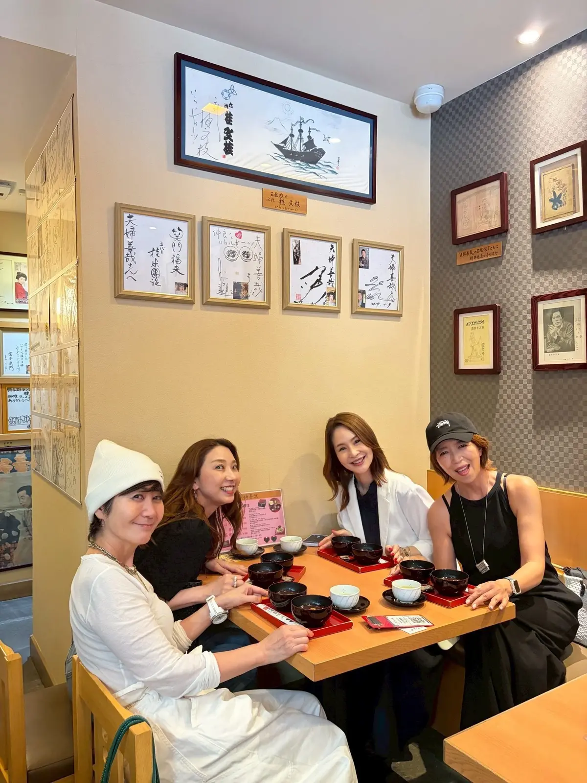 4人の女性がテーブルに座り、料理の器が並んだ和食レストランの内装。壁には額装された書や絵画が飾られ、チェック柄の壁と木製の家具が特徴。