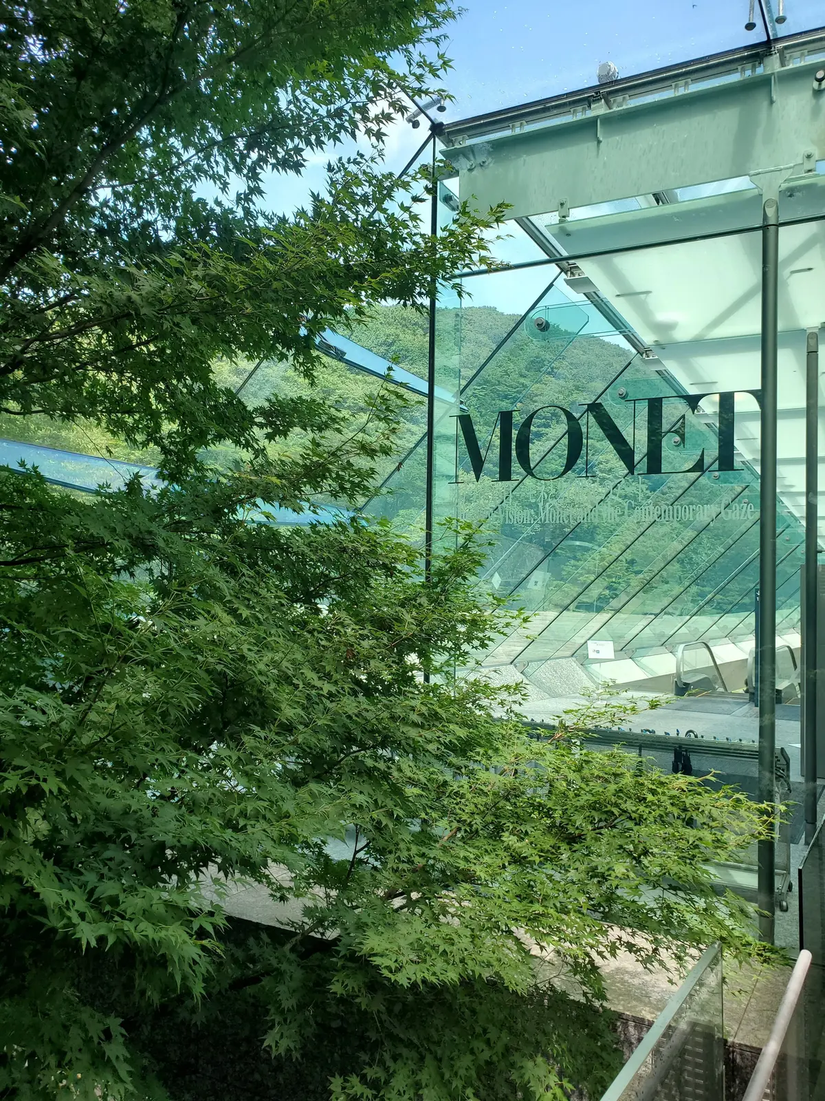 緑豊かな木々を背景に、ガラス張りの建物に「MONET」の文字が映し出されている。水辺の風景が建物越しに見える。