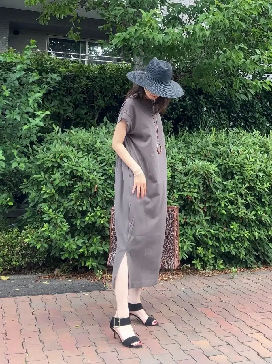 グレーのワンピースと帽子を着用した女性が、ブッシュの生い茂る庭を歩いています。彼女はレオパード柄のショルダーバッグを下げ、黒のハイヒールで歩いています。