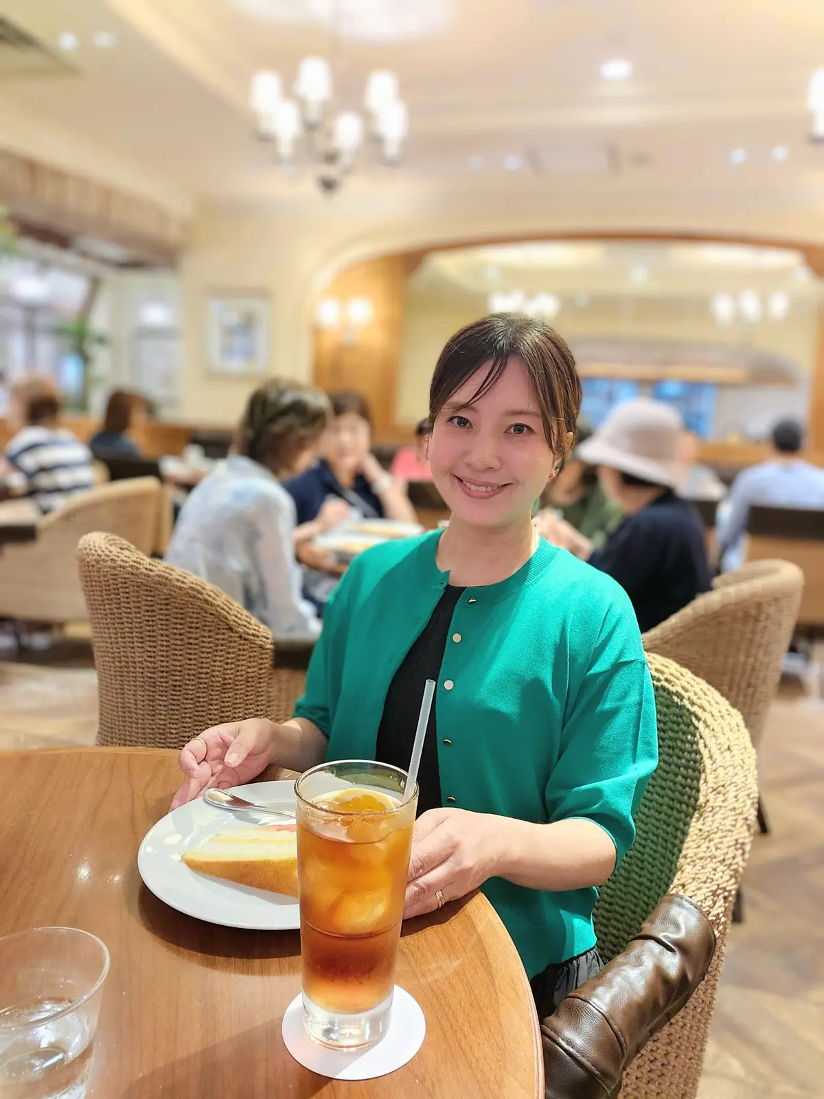 レストランで食事を楽しむ女性。緑色のカーディガンを着用し、笑顔でこちらを見ている。テーブルには飲み物とデザートが置かれている。背景には他の客席が見える。