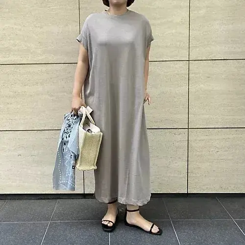 グレーのロングワンピースを着た女性が、壁に寄りかかりながら、トートバッグとスカーフを持っている