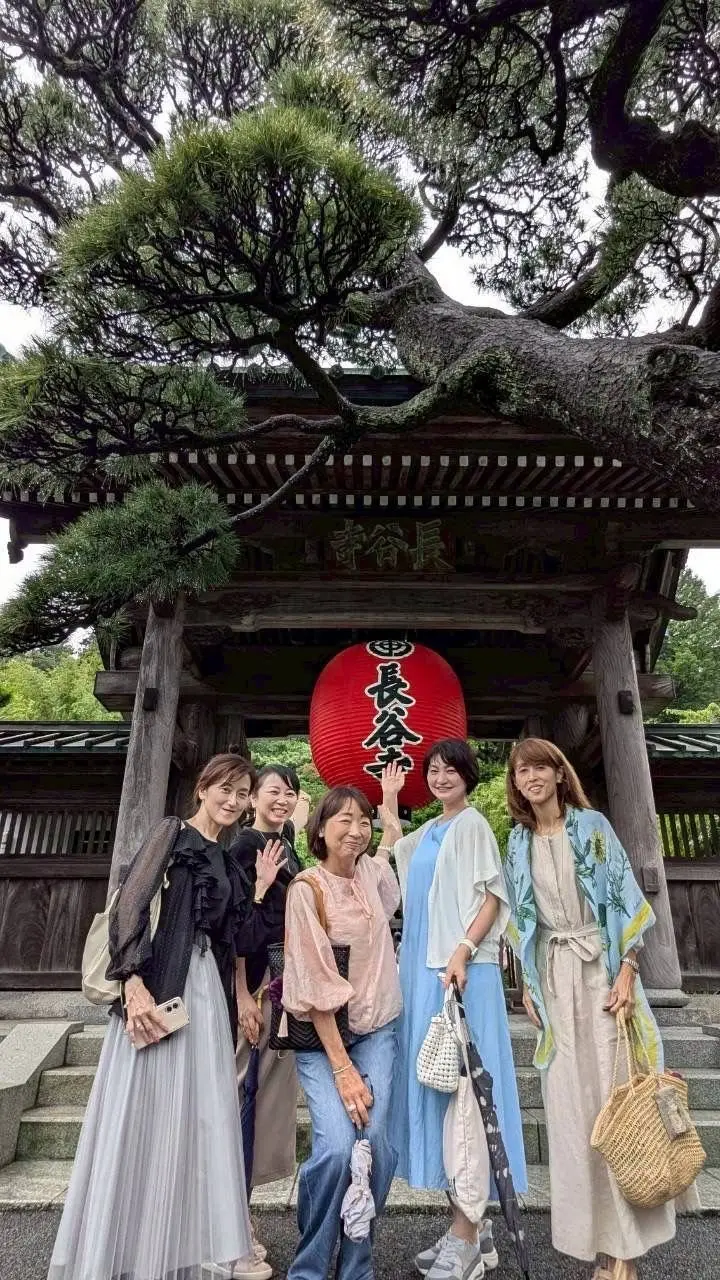 伝統的な寺院の鳥居前で、5人の女性が並んで写真を撮っている。中央に赤い提灯があり、背景には大きな木が広がっている。女性たちはそれぞれ異なる服装で、笑顔でカメラに向かって手を振っている。