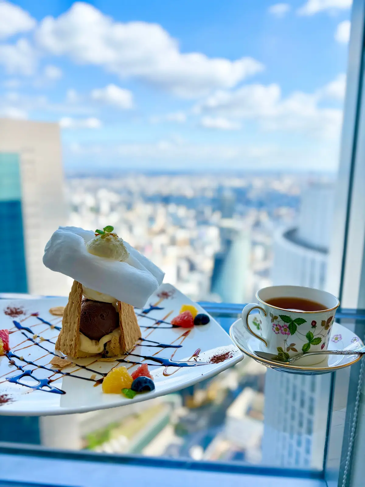 高層階の窓辺に置かれた、アイスクリームの器と紅茶のセット。アイスはワッフルコーンの中に盛り付けられ、メレンゲが添えられている。器の周りにはフルーツやチョコレートソースが飾り付けられている。背景には都市の景色と青空が広がる。