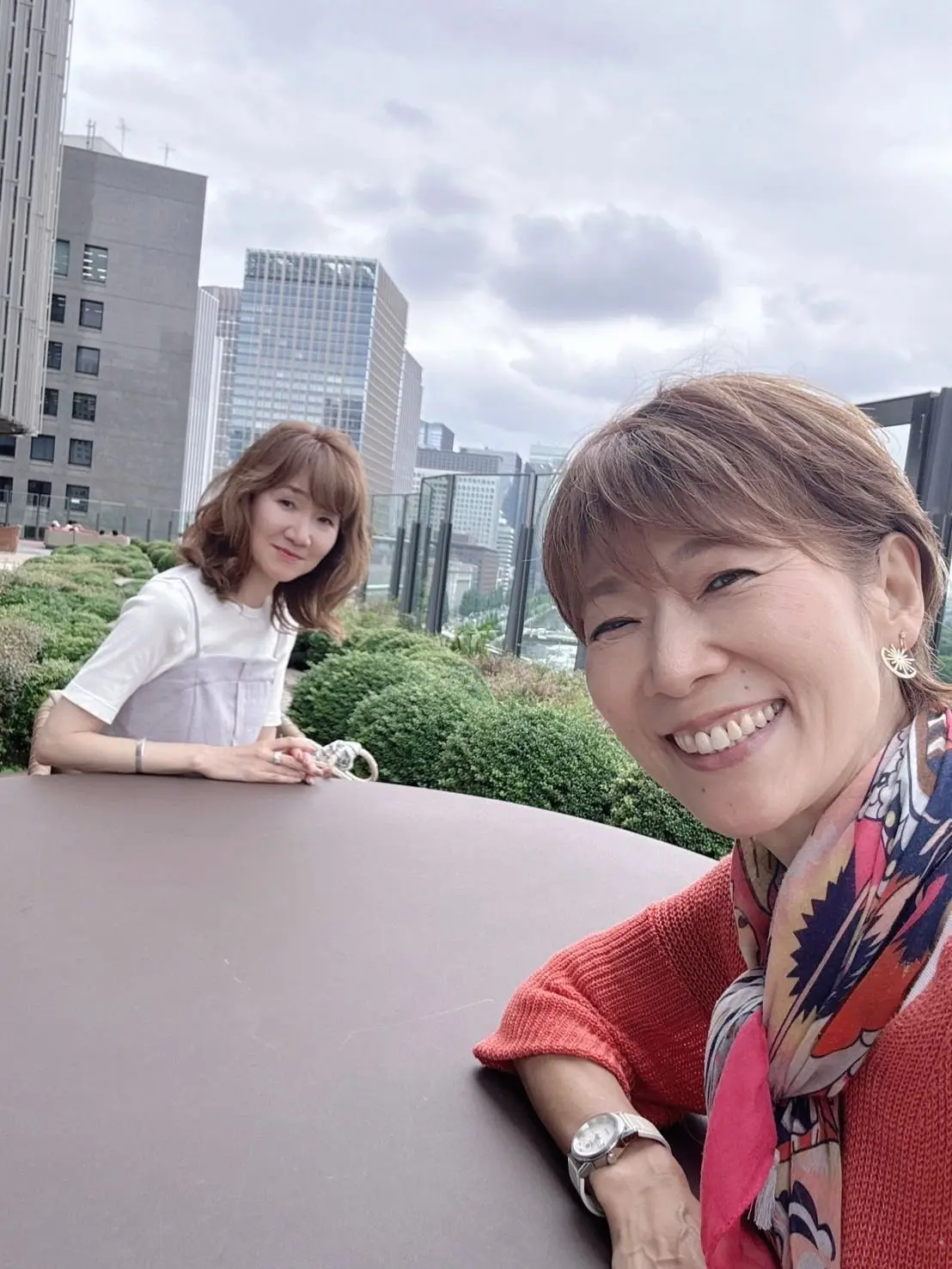女性二人が屋上テラスに座り、都市の高層ビルが背景に見える。左の女性は白い服で、右の女性は赤い服とスカーフを着て、笑顔でカメラに向かっている。