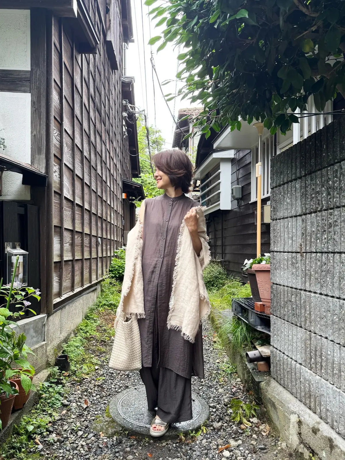 伝統的な木造家屋が並ぶ狭い路地で、女性が立っている。女性は茶色のワンピースとベージュのストールで、笑顔でカメラに向かって手をかざしている。路地には石の床、小さな植物、花鉢が置かれている。