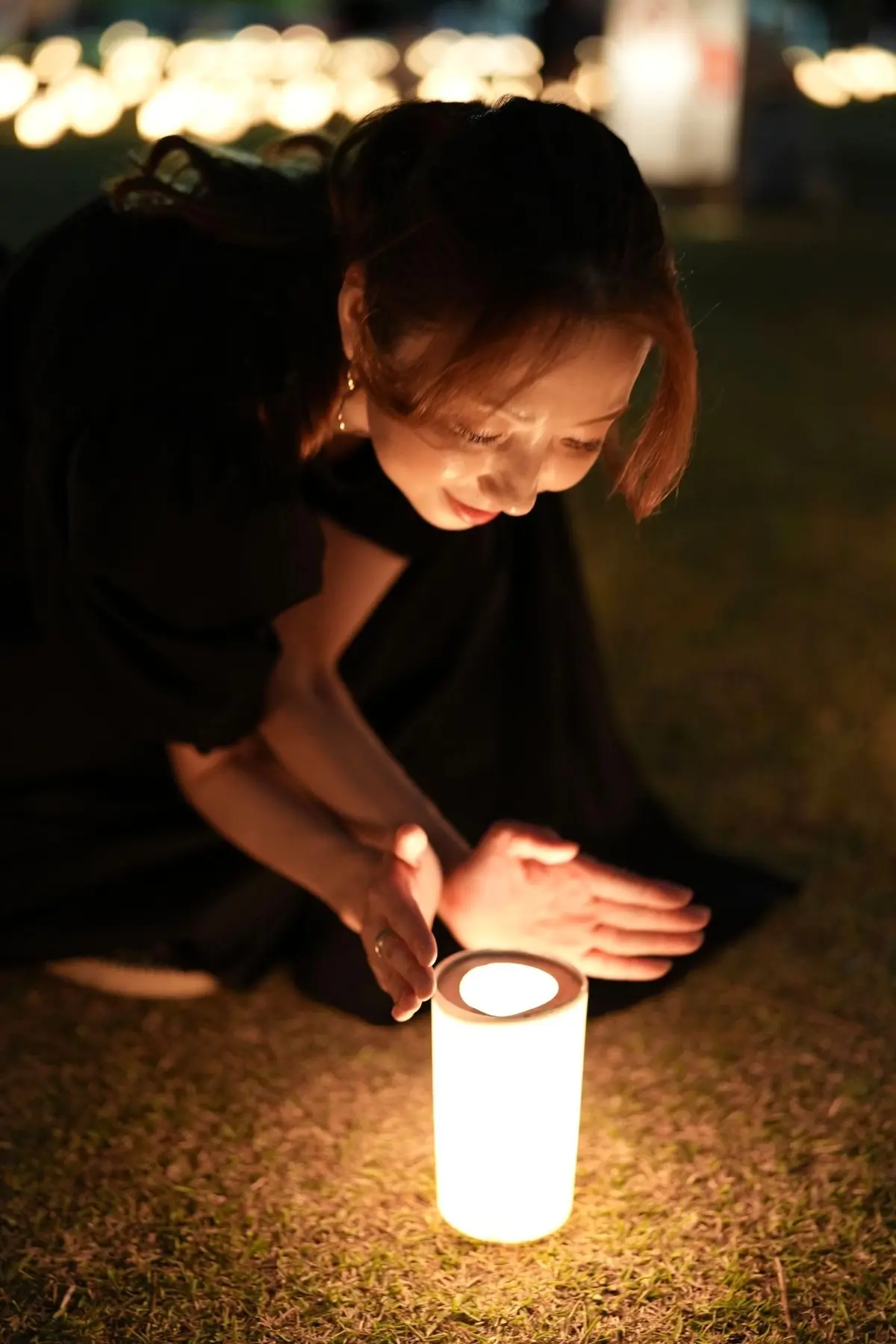 夜の芝生に膝をつき、若い女性が光る白いキャンドルを見つめている。黒い服を着ており、背景にはぼんやりと光るキャンドルが並んでいる。