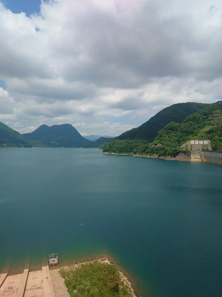 青緑の湖と緑豊かな山々が広がる風景。湖畔にはコンクリート構造のダムがあり、湖面には小型のボートが停泊している。空は部分的に雲が広がっている。