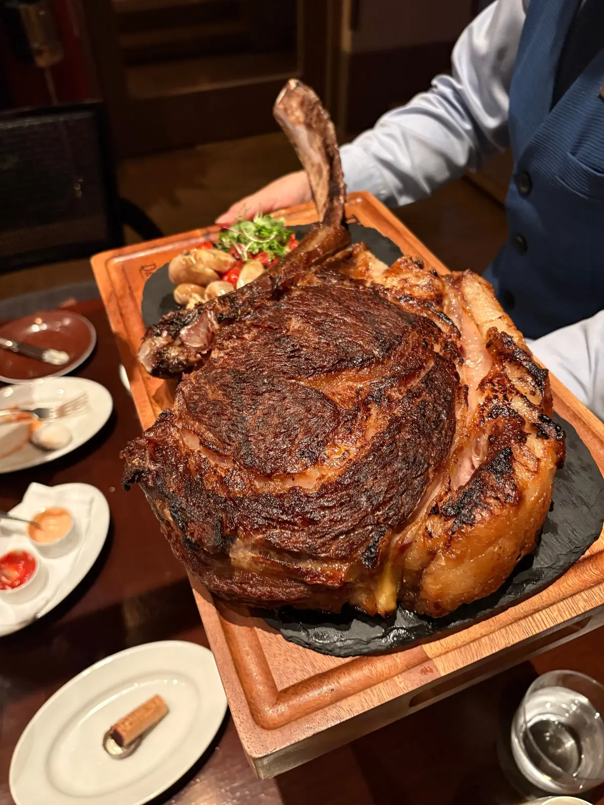 巨大なトマホークステーキが木製のボードに盛り付けられている。表面は焼き色で覆われ、骨付き肉が特徴的。周囲にはソースや付け合わせが置かれた白い皿が並んでいる。