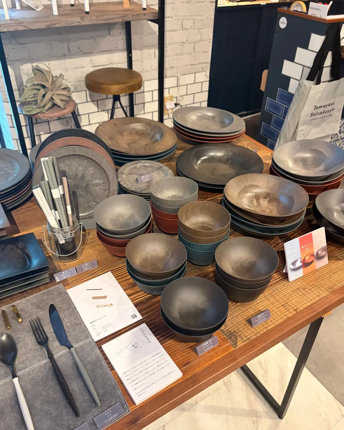 木製のテーブルに、陶器の食器やカトラリーが整然と並んでいる。様々な色と形の皿やボウルが置かれ、背景にはタイル張りの壁と白い紙袋が見える。