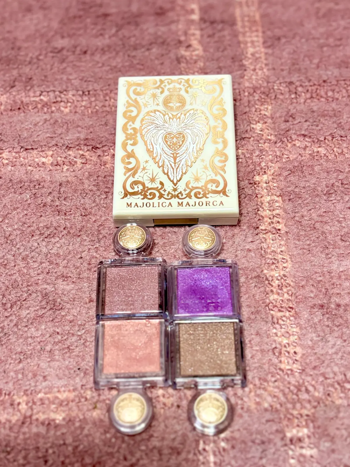 Majolica Majorcaのアイシャドウパレット。4色のコンパクトが並び、上から紫、ピンク、緑、茶色。コンパクトには金色の装飾が施され、背景はピンクがかったテクスチャ素材。