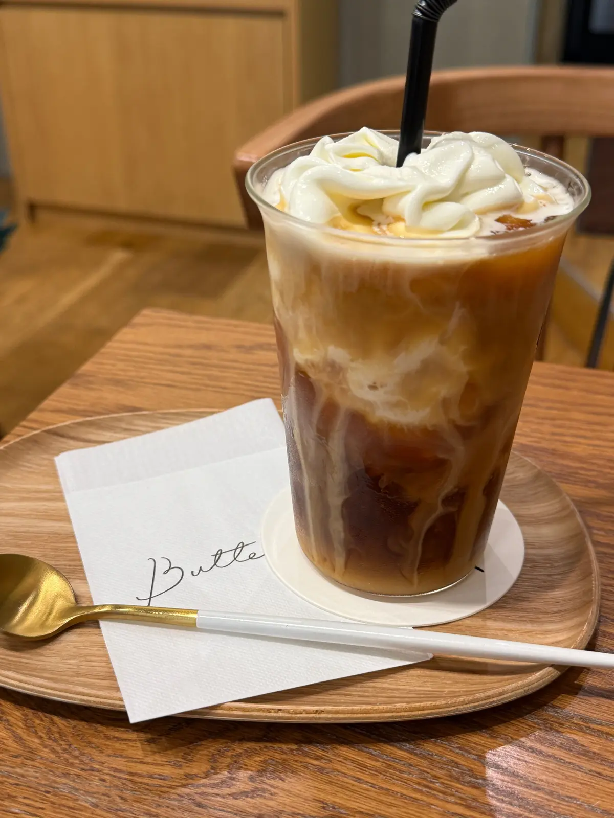木製のテーブルに置かれた、バターのロゴが入った白いコースターの上に、ホイップクリームが乗ったアイスコーヒー。黒いストローとスプーンが添えられている。
