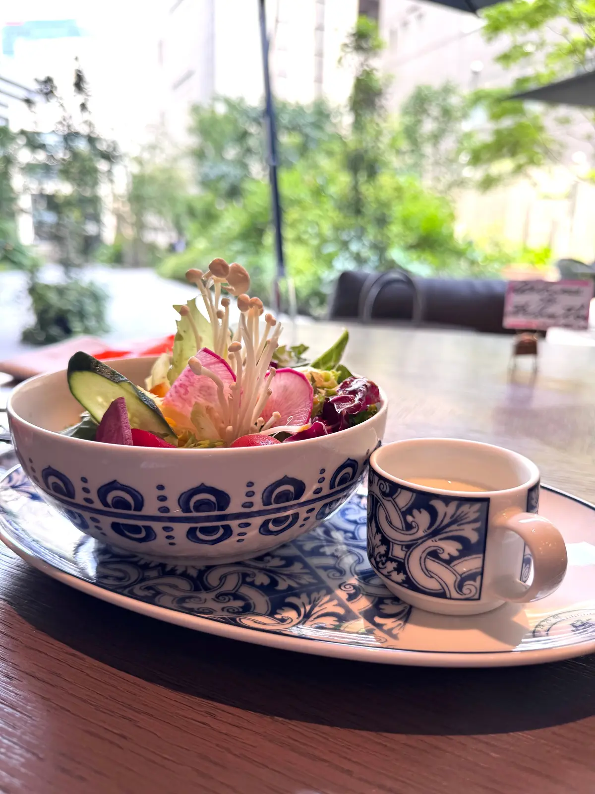 青と白の模様のある陶器のボウルに、きゅうり、レタス、ラディッシュ、花のような装飾が添えられたサラダが盛り付けられ、隣には同じ模様のマグカップにコーヒーが置かれている。