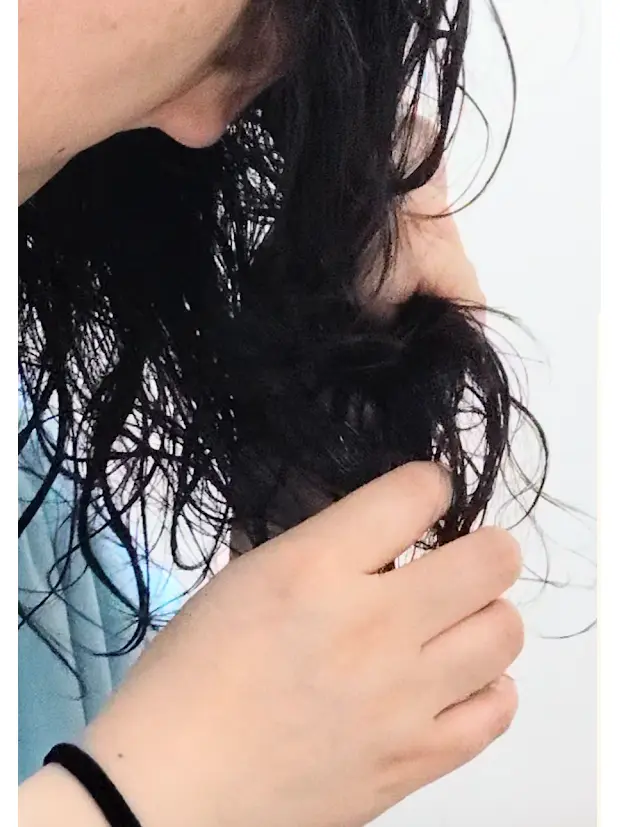 黒い髪の女性が、手で髪を掴んでいるアップ写真。白いシャツを着用し、黒いリストバンドをしている。背景は白。