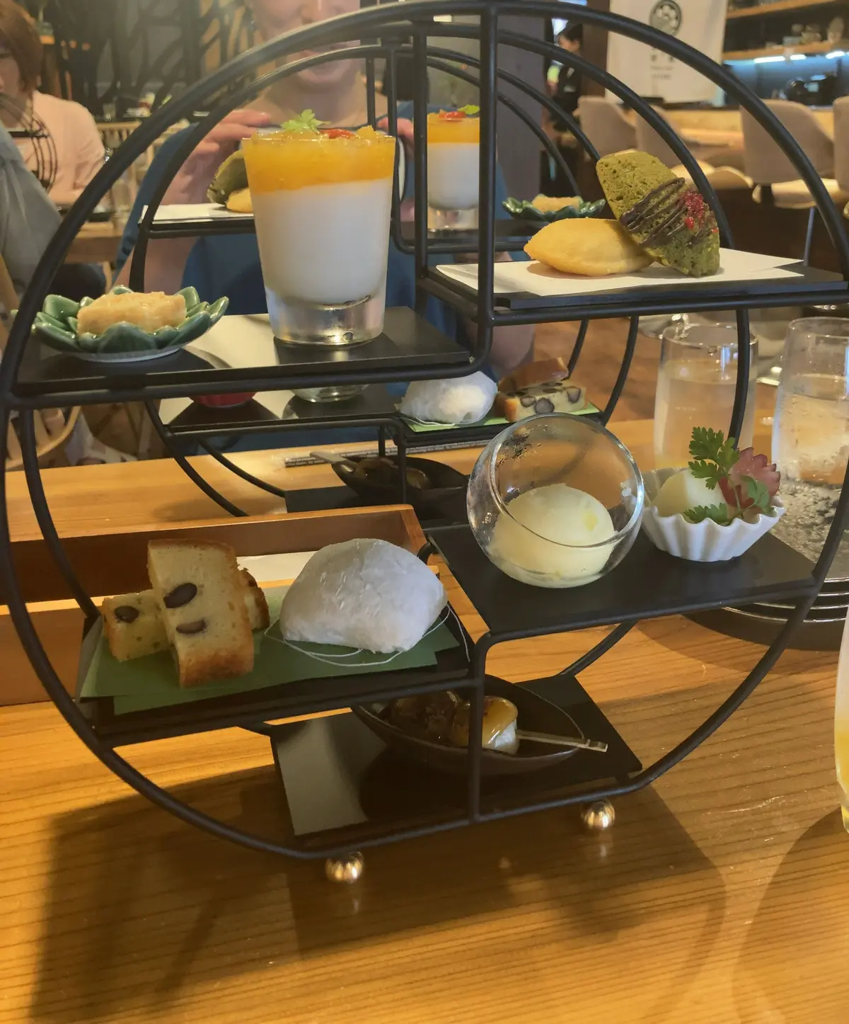 円形のスタンドに、層状のデザート、緑茶のクッキー、フルーツの盛り合わせ、パン、クリームなど、さまざまなスイーツとドリンクが並んでいる。