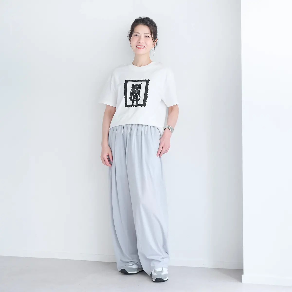 白いTシャツに黒いイラストがプリントされた女性。グレーのワイドパンツを着用し、白いスニーカーを履いている。背景は白い壁。