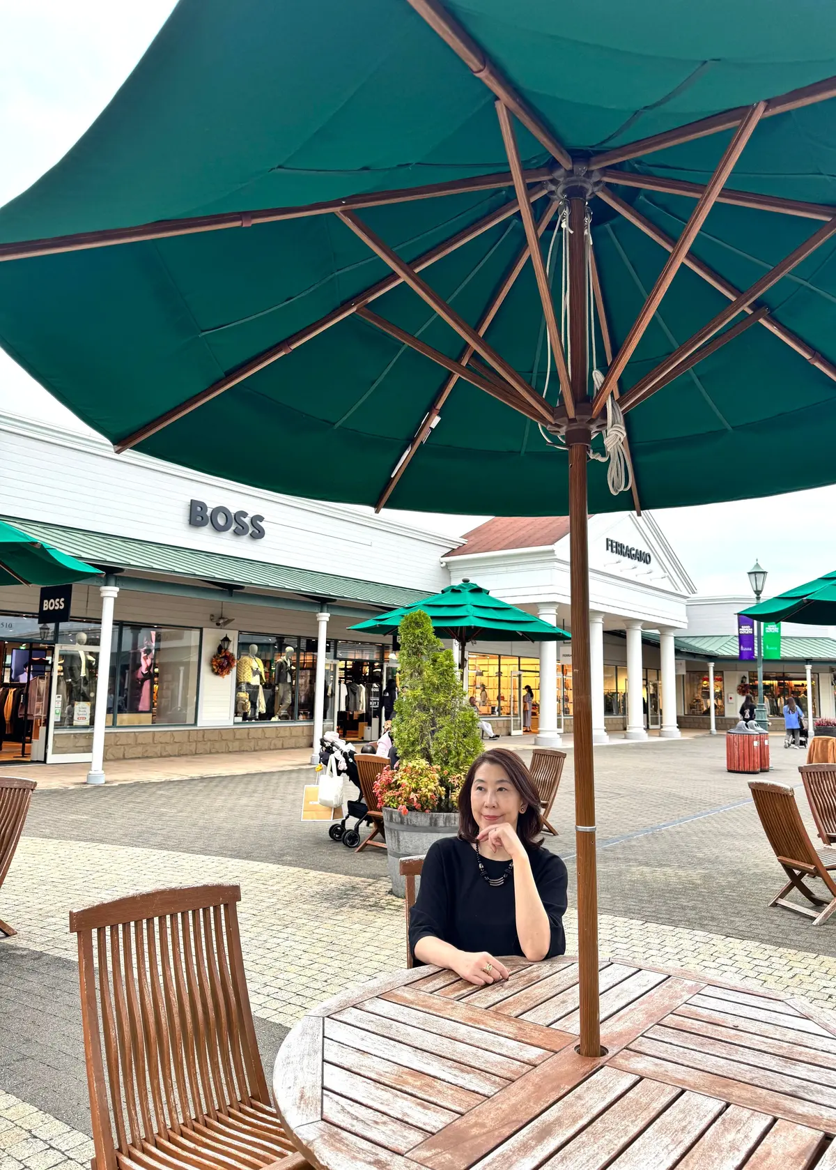 緑色のパラソル下に座る女性。背景には、BOSSやFERRAGAMOなどのブランド店が並ぶアウトレットモール。木製のテーブルと椅子が配置され、植栽された常緑樹が見える。