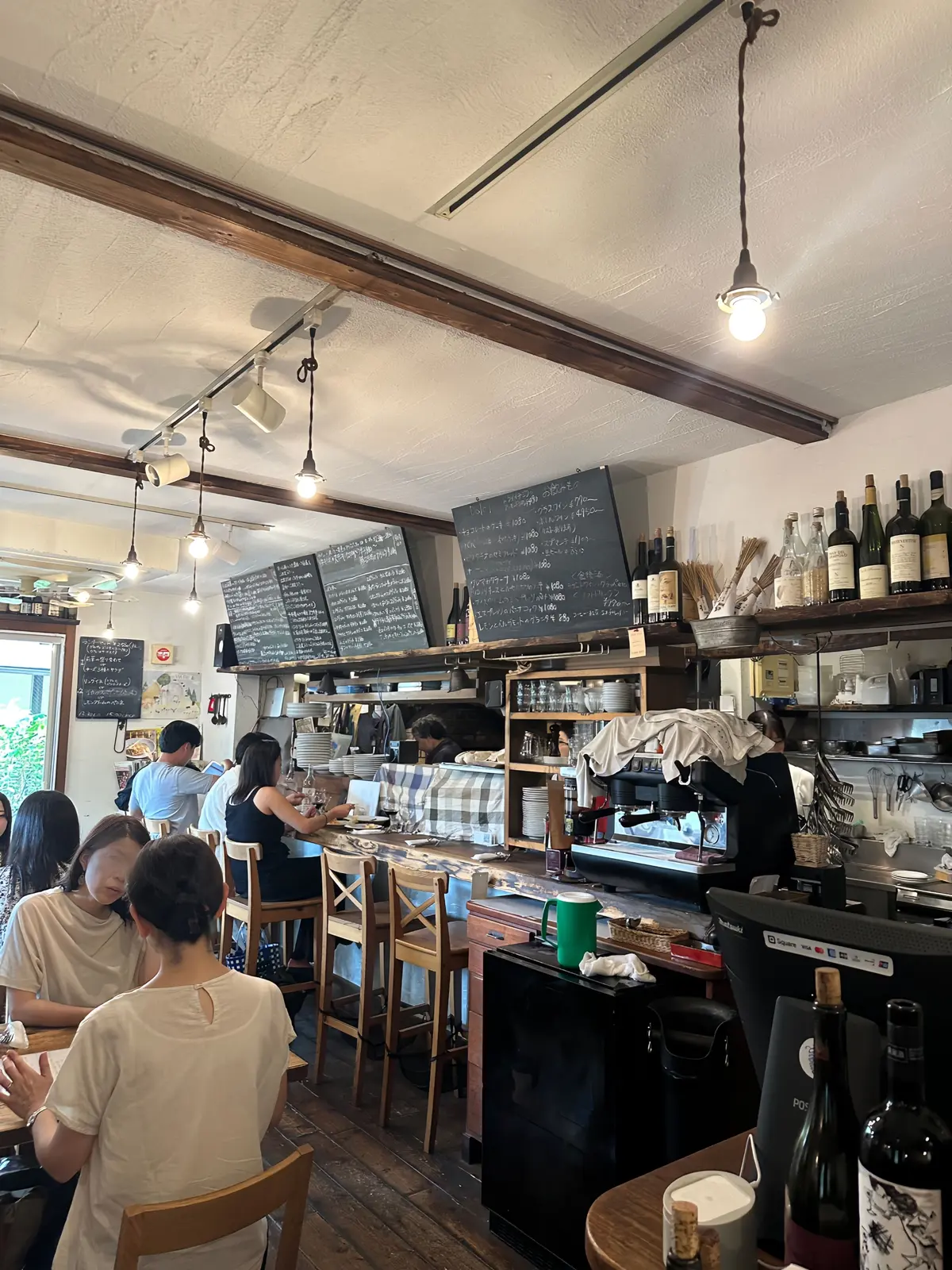 木目調の梁が走るカフェの店内。手書きのメニューが並び、カウンターでは店員が作業中。テーブル席には客が座り、落ち着いた雰囲気。