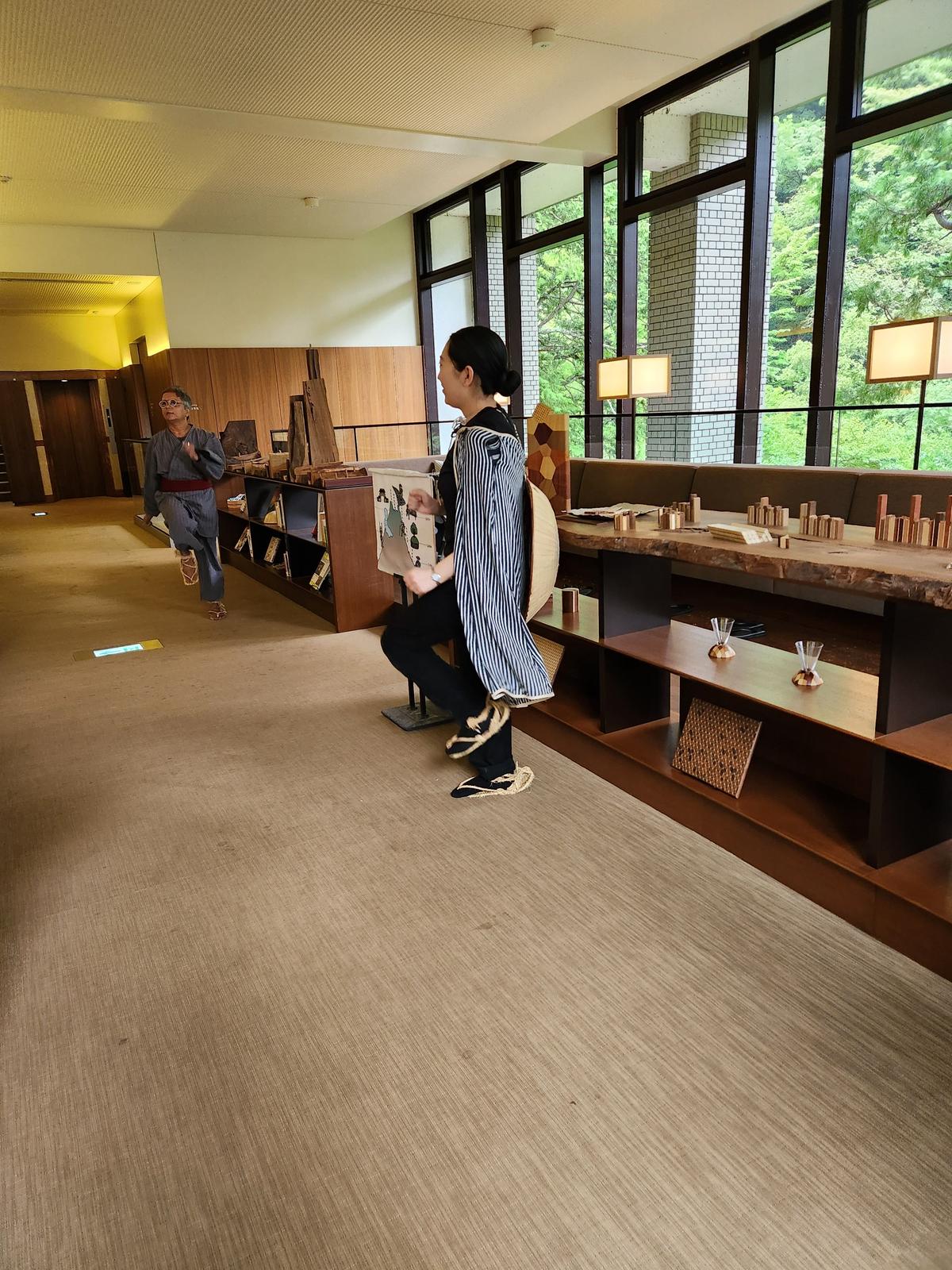 広い部屋に、木製の棚と窓、そして2人の人がいる。左の人は赤い着物姿で、右の人はストライプの服を着て座っている。窓の外には緑豊かな木々が見える。