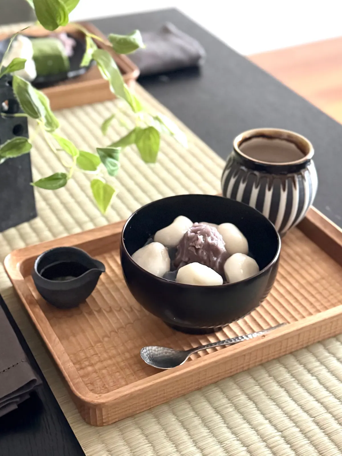 木製トレイに黒いボウルに入った白い豆と紫の餡、小さな黒い容器、黒と白の模様の陶器カップ、木製のスプーンが置かれています。