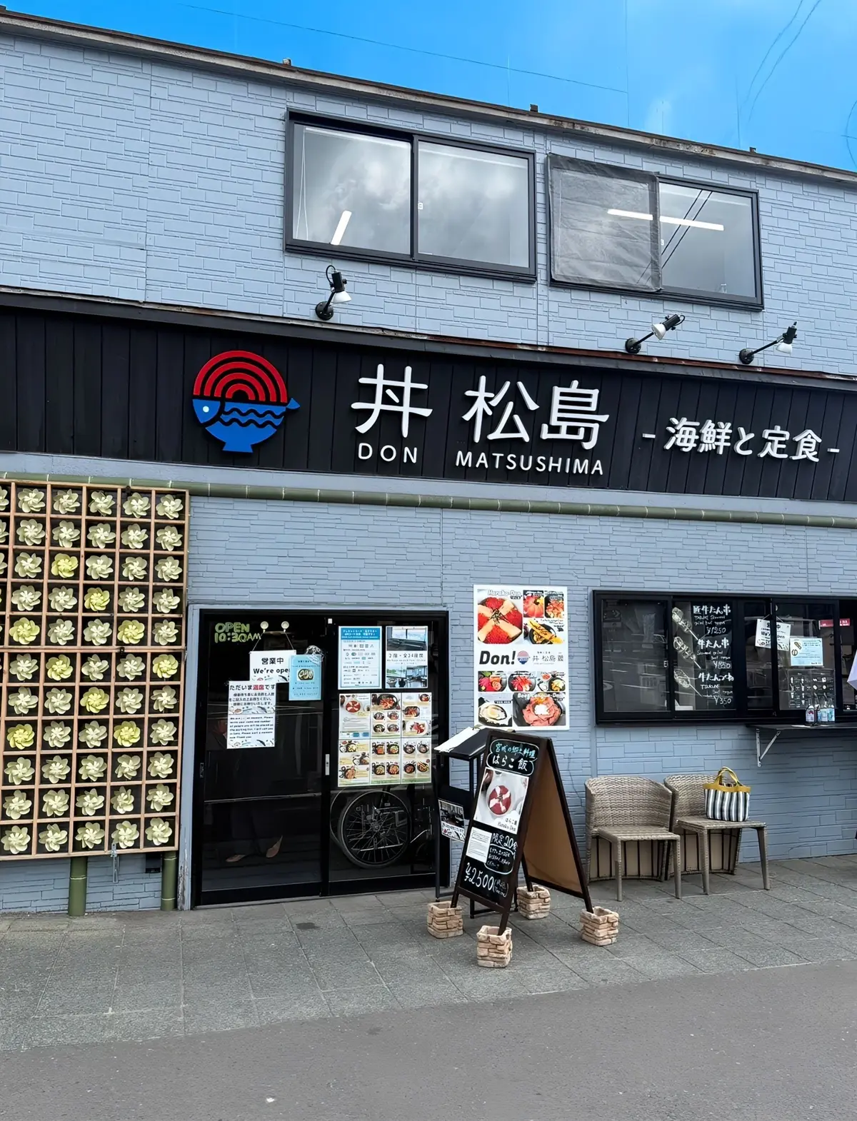 灰色の外壁のレストラン「丼 まつしま」の正面。窓にメニューが貼られ、入り口前に看板と椅子が置かれている。ロゴは青地に白い波のデザイン。