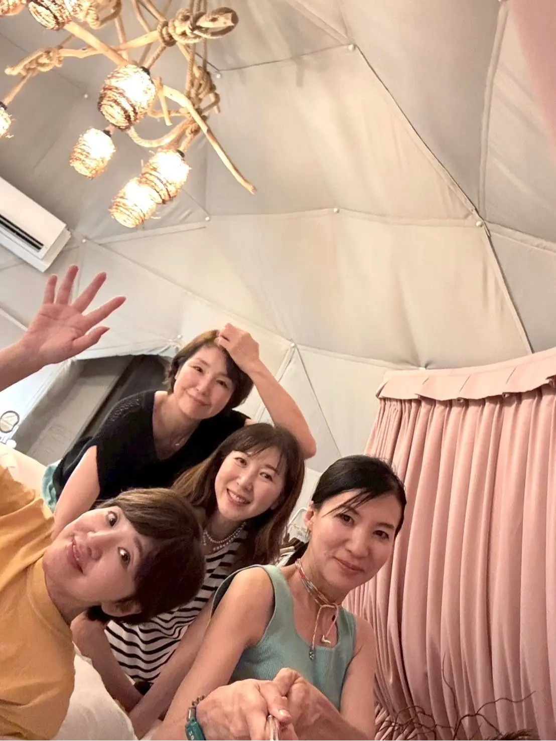 テント内にて、4人の女性が寄り添って笑顔でポーズ。手前から黄色、縞模様、薄緑、黒の服を着た女性が前列に座り、後列に手で髪を整える女性がいる。天井には複数の電球が取り付けられた木製の照明が吊るされている。