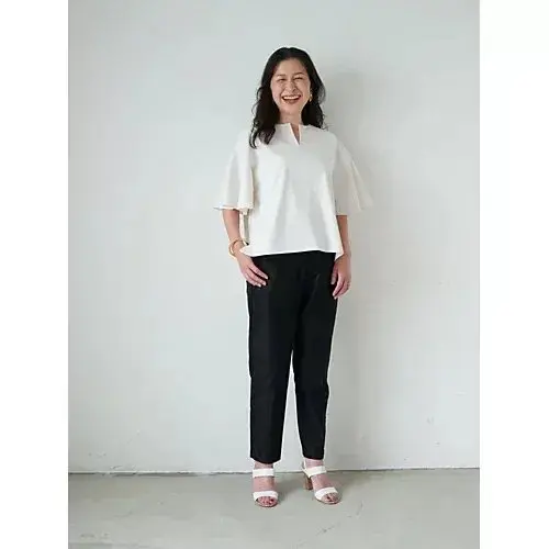 白い襟ぐりのトップスに黒いパンツを着用した女性が、白い背景の前で微笑んでいる全身写真。金色のイヤリングとブレスレットを身に着けている。