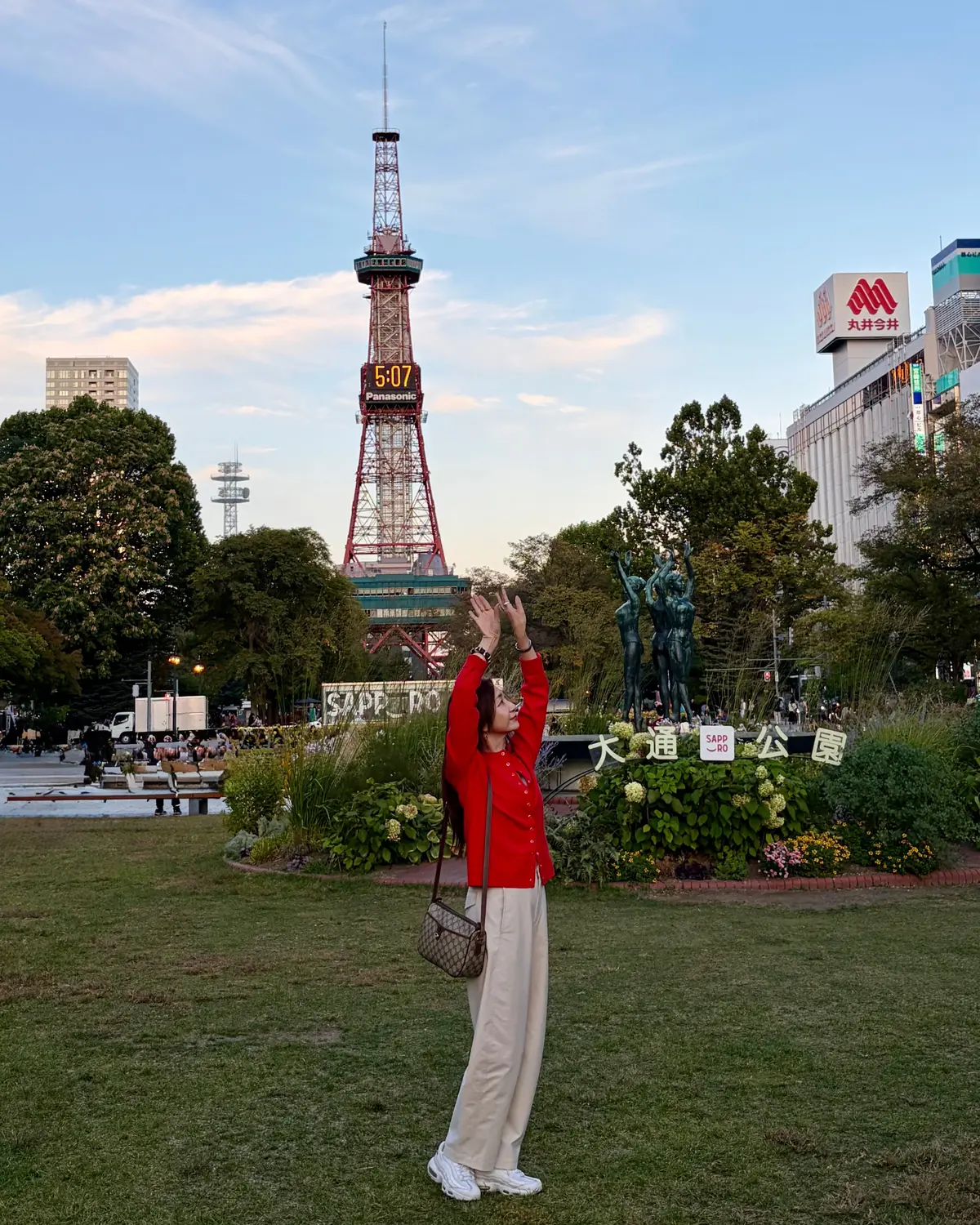 赤いトップスとベージュのパンツを着た女性が、芝生の上で手を挙げている。背景には、さっぽろテレビ塔と周囲の建物、青空が広がる。