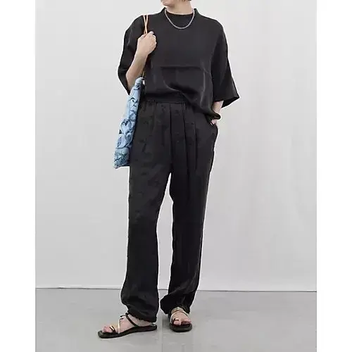 黒いサテン素材のトップスとパンツを合わせた全身コーディネート。シルバーのネックレスとサンダルを着用。背景には青色のプリントバッグ。