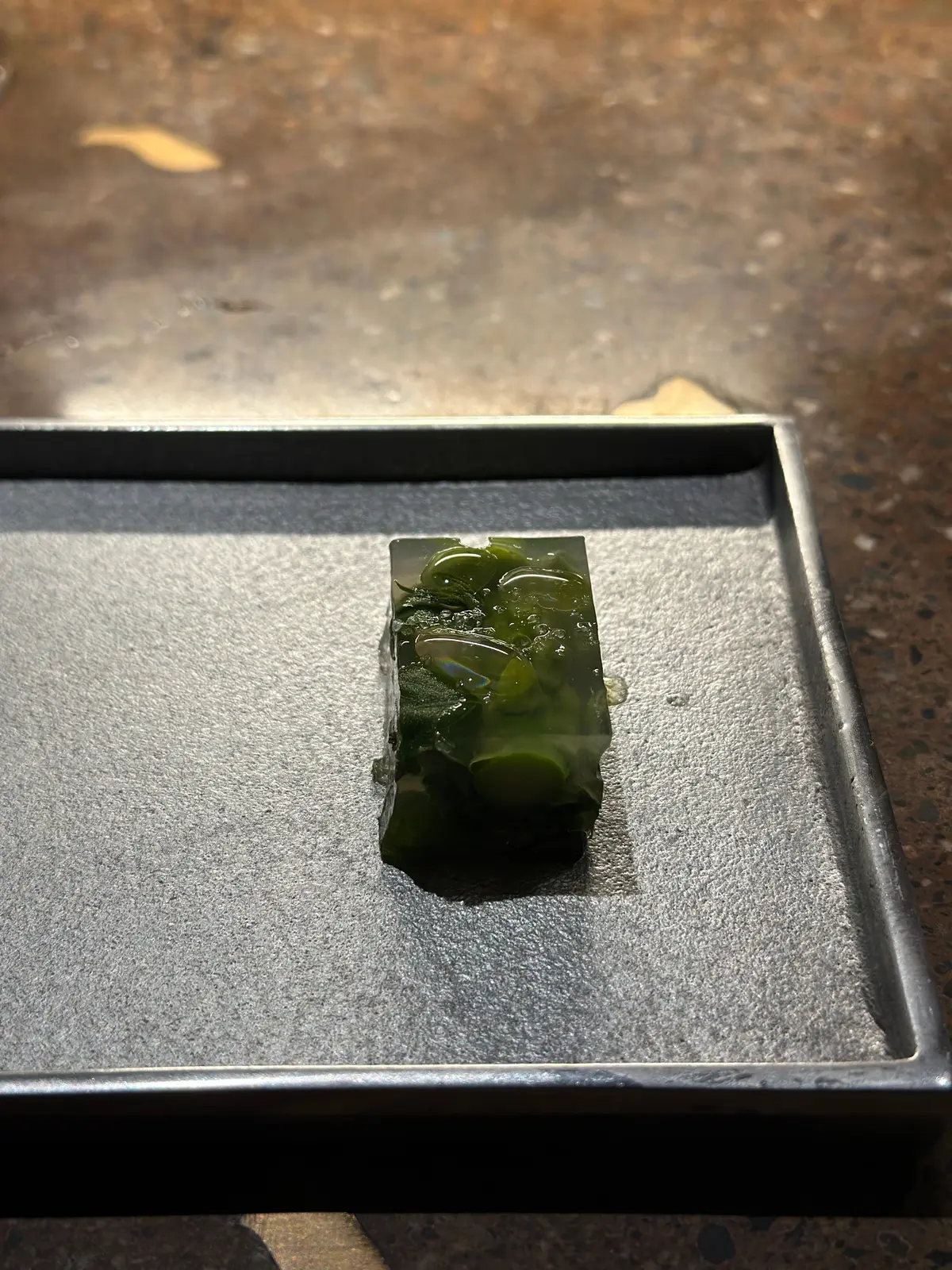 灰色のトレイに置かれた、濃い緑色の長方形の翡翠彫刻。龍のような生き物が彫り込まれており、表面には苔のような模様がある。