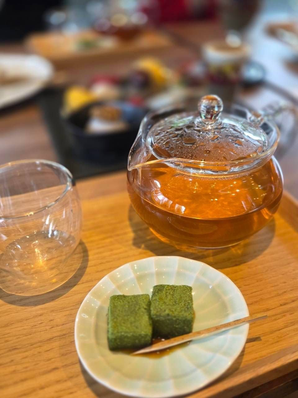 木製のトレーに置かれた、透明なティーポットに入ったお茶と、淡い緑色の和菓子２つ。ガラス製のグラスと、背景に食器が置かれている。