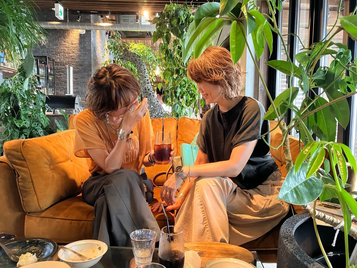 カジュアルな服装の2人の若い男女が、緑豊かな植物に囲まれたオレンジ色のソファに座り、テーブルを挟んで楽しそうに会話している様子。テーブルにはグラスや料理が置かれている。